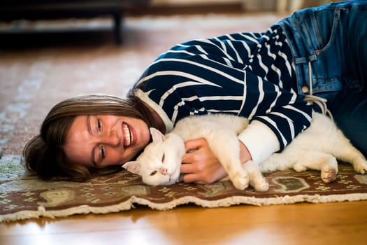 A young cute girl lies on the carpet of the room in an embrace with her white fluffy pet, a woman in a striped sweater smiles and hugs her purring best friend cat, the owner takes care of her kitty.