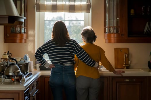 The daughter hugs her elderly mother, the granddaughter and her grandmother enjoy life, look out the window at a beautiful view from their cozy home, spend time preparing together in the kitchen.