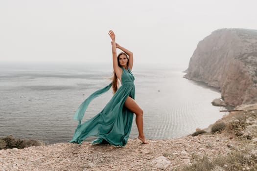 Side view a Young beautiful sensual woman in a mint long dress posing on a volcanic rock high above the sea during sunset. Girl on the nature on overcast sky background. Fashion photo