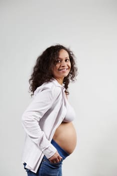Studio shot of a curly haired mixed race happy pregnant woman 30s, smiling cutely looking aside, posing with hands in jeans pockets and bare belly, over isolated white background. Pregnancy fashion.