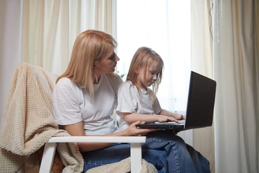 Beautiful young woman and her little cute daughter are using laptop at home. Enjoying spending time together with Internet and modern technologies