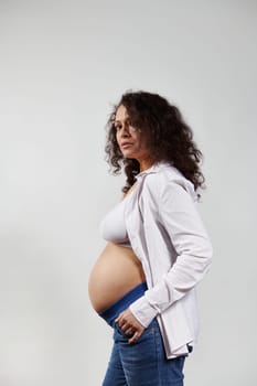 Vertical studio portrait of curly haired brunette pregnant woman, wearing blue jeans and unbuttoned white shirt, posing bare belly over isolated white background. Happy pregnancy and maternity concept