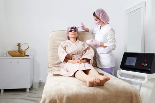 Cosmetologist applying black mask on the face of a beautiful woman for carbon peeling in beauty salon