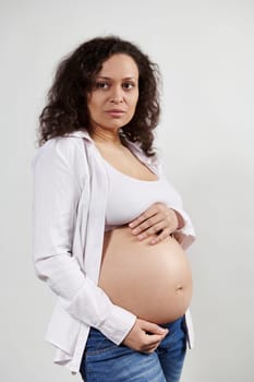 Vertical studio portrait of beautiful pregnant Hispanic woman with curly dark hair, expecting a baby. Happy future mom in 30 weeks of pregnancy, posing with hands on naked belly over white background.