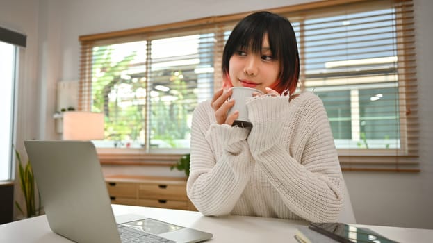 Portrait of young Asian woman drinking coffee and using laptop at home. Freelance, e-learning and remote working concept.