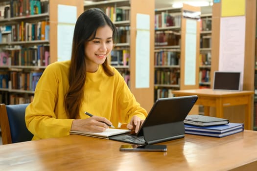 Happy asian student girl attending online class, studying in library with digital tablet. Education and technology concept.