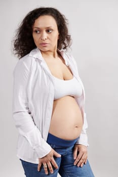 Vertical studio portrait of curly haired multi ethnic confident serene pregnant woman, wearing unbuttoned white shirt and blue denim, looking aside on isolated white background. Pregnancy. Maternity