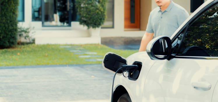 Progressive asian man install cable plug to his electric car with home charging station in the backyard. Concept use of electric vehicles in a progressive lifestyle contributes to clean environment.