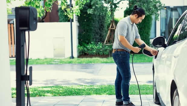 Progressive asian man install cable plug to his electric car with home charging station in the backyard. Concept use of electric vehicles in a progressive lifestyle contributes to clean environment.