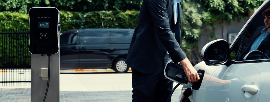 Closeup progressive black suit businessman insert EV charger plug from public charging station to his electric car. Eco friendly electric rechargeable car with zero CO2 emission.