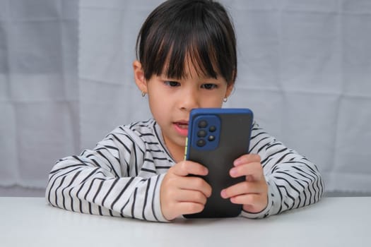 Cute little girl using smartphone and smiling while sitting at table in house. Happy Asian girl holding phone in hands, online learning or homeschooling, listening to music or playing games.