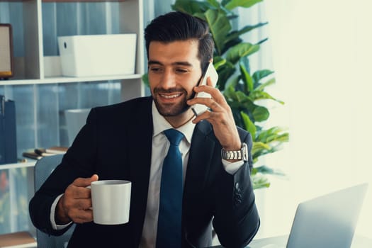 Businessman working in modern office workspace with cup of coffee in his hand while answering phone call making sales calls or managing employee. Fervent