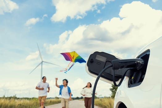 Concept of progressive happy family holding windmill toy and relax at wind farm with electric vehicle. Electric vehicle driven by clean renewable energy from wind turbine generator to charger station.