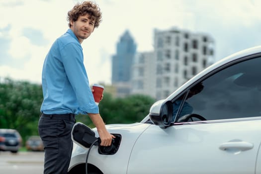 Progressive eco-friendly concept of parking EV car at public electric-powered charging station in city with blur background of businessman leaning on recharging-electric vehicle with coffee.