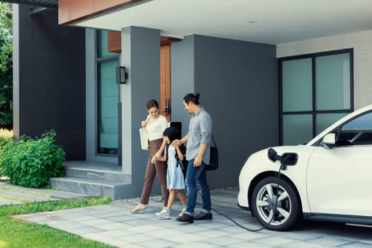 Progressive young parents and daughter with electric vehicle and home charging station. Green and clean energy from electric vehicles for healthy environment. Eco power from renewable source at home.