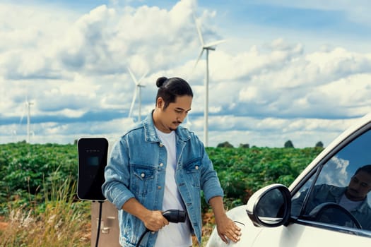 Progressive man with his electric car, EV car recharging energy from charging station on green field with wind turbine as concept of future sustainable energy. Electric vehicle with energy generator.