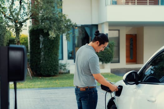 Progressive asian man install cable plug to his electric car with home charging station in the backyard. Concept use of electric vehicles in a progressive lifestyle contributes to clean environment.