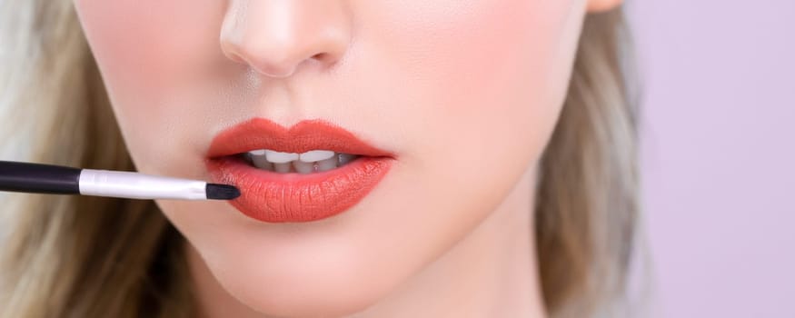 Closeup beautiful young woman with flawless healthy skin and natural makeup putting alluring fashion glossy red lipstick on her lip with lip brush in pink isolated background.