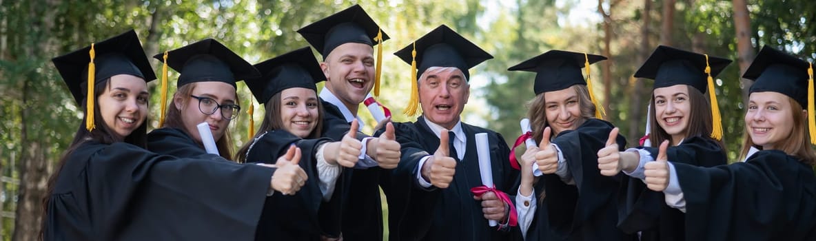 A group of graduates in robes give a thumbs up outdoors. Elderly student. Widescreen