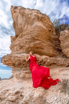 A woman in a red silk dress stands by the ocean, with mountains in the background, as her dress sways in the breeze