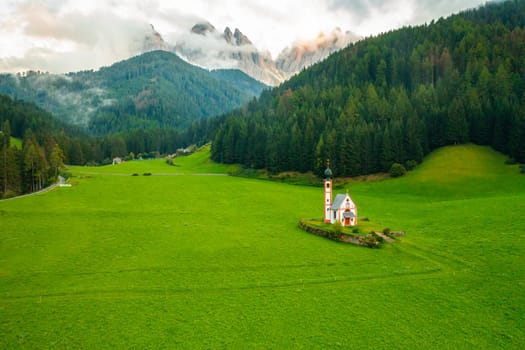 Aerial view of the Church of St. Johann against the Geisler peaks covered with clouds in Santa Maddalena village, Val Di Funes, Italy.