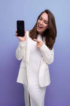 smiling young brunette with long hair lady in white dress shows smartphone screen with mockup.