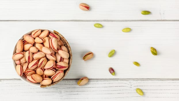 Tabletop view, small bowl with red Turkish pistachios, more nuts scattered on white board desk.