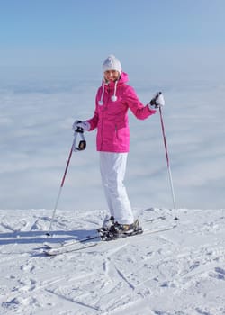 Woman in pink jacket, ski poles and skis, standing at the edge of hill, only clouds below, looking back, smiling