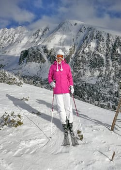 Young woman in pink jacket,  ski, boots, poles, gloves and hat, posing on the piste, snow covered mountain behind her during sunny day.