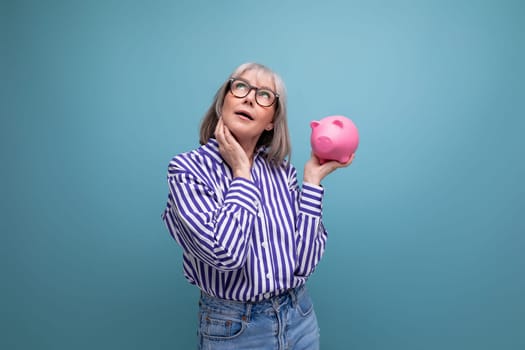 pensive 60s woman with gray hair holding piggy bank thinks about insurance on bright studio background.