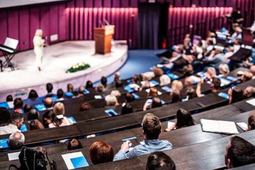 Business and entrepreneurship symposium. Female speaker giving a talk at business meeting. Audience in conference hall. Rear view of unrecognized participant in audience.