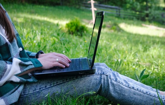 Young woman's legs on the green grass with open laptop. Girl's hands on keyboard. Distance learning concept. Happy hipster young woman working on laptop in park. download image