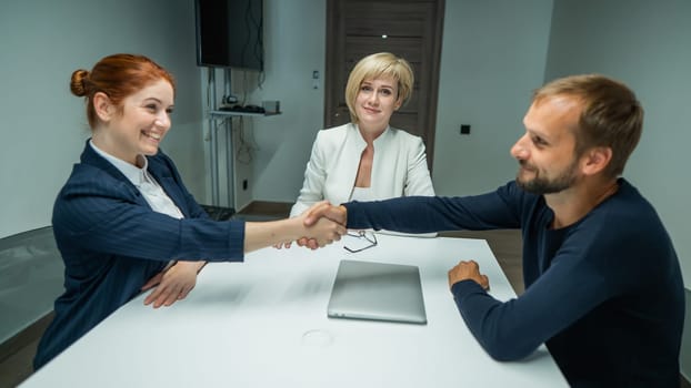 Blond, red-haired woman and bearded man dressed in suits in the office. Business people shake hands making a deal in a conference room