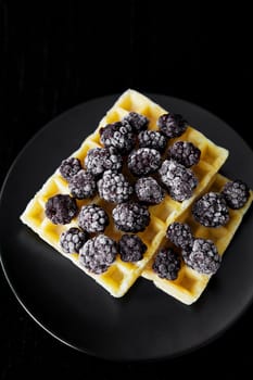 Belgian wafers with frozen blackberries on a black background, close-up.