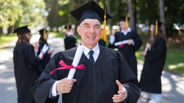 A group of graduates in robes outdoors. An elderly student rejoices at receiving a diploma