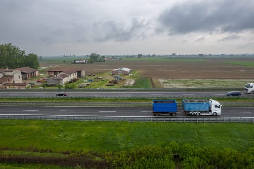 Piacenza, Italy - April 2023 Cargo trucks and cars in the motorway a1 autostrada del sole aerial point of view in between padain plain rural fields