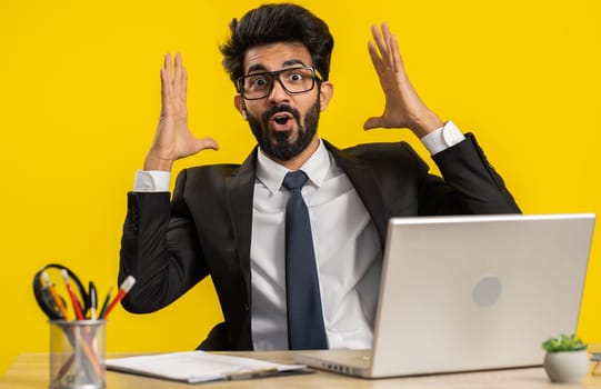 Excited amazed indian businessman touching head, showing explosion, looking worried shocked, professional burnout at office workplace. Hindu man in formal suit working on laptop on yellow background