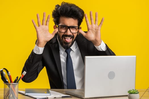 Indian businessman use laptop computer, making funny face, fooling around, putting his tongue out, remote working disrespecting someone at office desk workplace. Freelancer man on yellow background