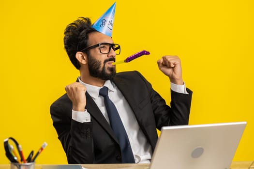 Happy indian businessman wearing birthday hat cone blow pipe celebrating success win victory at office workplace desk. Excited hindu man in formal suit working on laptop isolated on yellow background
