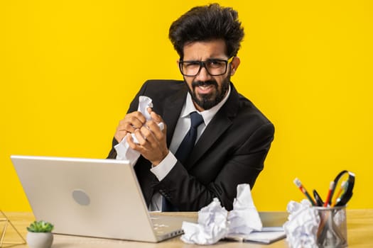 Angry furious indian man working at office throwing crumpled paper, having nervous breakdown at work, migaine, headache, stress management, mental distress problems, losing temper, reaction on failure