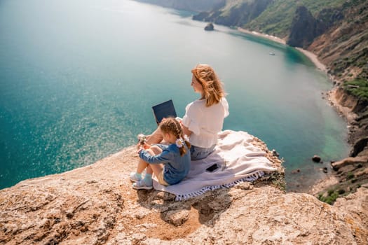 Freelance woman with her daughter working on a laptop by the sea, typing on the keyboard, enjoying the beautiful view, highlighting the idea of remote work