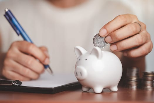 Asian women's hand saving a coin into piggy bank, planning and writing information on paper with blurred coins and money on the wooden table for investment, business, finance and saving money concept.