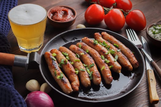 Delicious sausages cooked in a frying pan with vegetables, various sauces and a glass of beer on a wooden table.