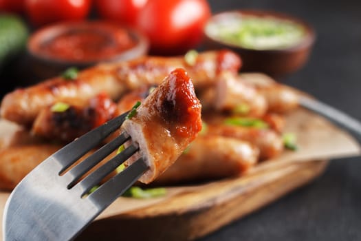 A piece of sausage on a fork against the background of sausages with vegetables and various sauces on the table.