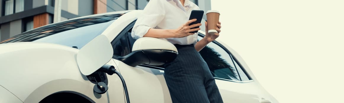 Focus businessman using phone, leaning on electric vehicle, holding coffee with blurred city residential condo buildings in background as progressive lifestyle by renewable and sustainable EV car.