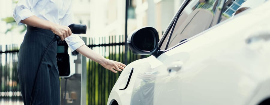 Progressive businesswoman insert charger plug from charging station to her electric vehicle with apartment condo building in background. Eco friendly rechargeable car powered by sustainable energy.