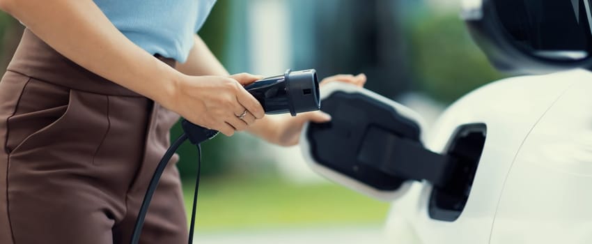 Closeup progressive woman install cable plug to her electric car with home charging station. Concept of the use of electric vehicles in a progressive lifestyle contributes to clean environment.