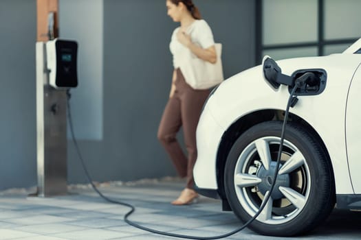 Focus image of electric vehicle recharging battery at home charging station with blurred woman walking in the background. Progressive concept of green energy technology applied in daily lifestyle.