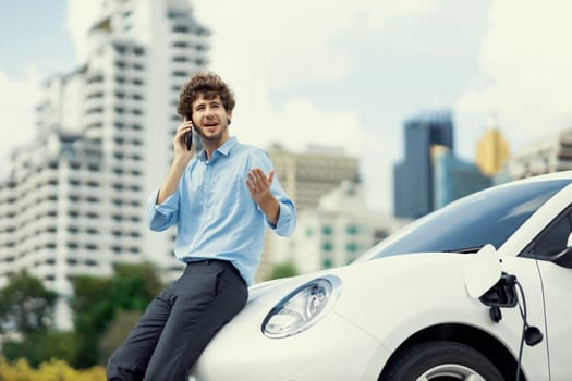 Progressive businessman talking on the phone, leaning on electric car recharging with public EV charging station, apartment condo residential building on the background as green city lifestyle.