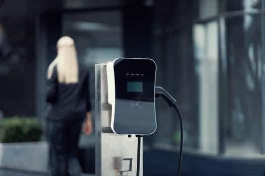 Focus public EV charging station for electric car with blurred progressive businesswoman walking in background. Electric car driven by clean and sustainable energy for ecological concern.